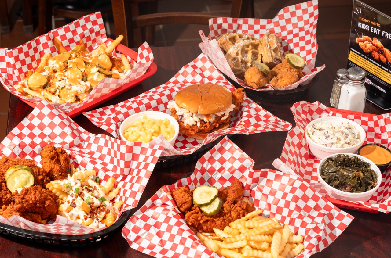 Overhead spread of Bold Birds Nashville hot chicken — loaded fries, hot chicken sandwich, chicken and waffles, tenders, collards and potato salad